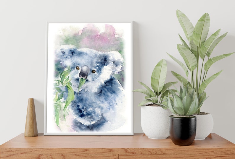 May include: Watercolour painting of a koala bear eating eucalyptus leaves. The artwork features shades of blue and grey, with a touch of pink in the background. The framed print is displayed on a wooden surface, with potted plants in the background.