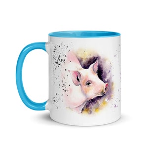 May include: A white ceramic mug with a light blue rim and handle. The mug features a watercolor illustration of a pink pig with a light brown background and black splatters.