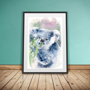 May include: Watercolour painting of a koala bear eating eucalyptus leaves. The artwork features shades of blue, grey, and green, with a soft, artistic style. The framed print is displayed against a teal wall and wooden floor.