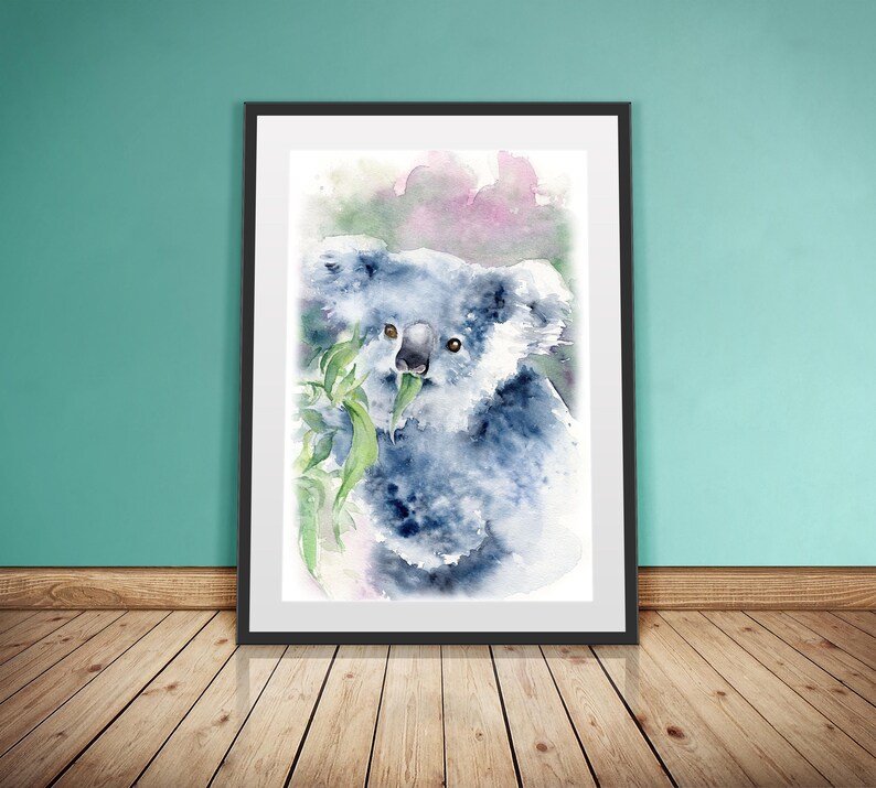 May include: Watercolour painting of a koala bear eating eucalyptus leaves. The artwork features shades of blue, grey, and green, with a soft, artistic style. The framed print is displayed against a teal wall and wooden floor.