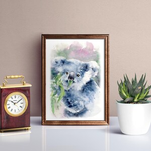 May include: Watercolour painting of a koala bear eating eucalyptus leaves, framed in a brown wooden frame. A small, decorative clock with a gold handle and a white-faced clock is on the left. A potted succulent is on the right.