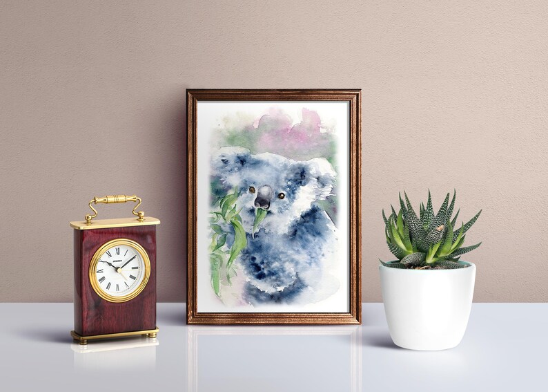 May include: Watercolour painting of a koala bear eating eucalyptus leaves, framed in a brown wooden frame. A small, decorative clock with a gold handle and a white-faced clock is on the left. A potted succulent is on the right.