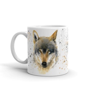 K&ouml;nnte beinhalten: Wei&szlig;e Keramik-Tasse mit einer Aquarell-Illustration eines Wolfsgesichts. Der Wolf hat graues und braunes Fell und gelbe Augen. Goldene und schwarze Spritzer umgeben das Portr&auml;t. Die Tasse hat einen wei&szlig;en Henkel.