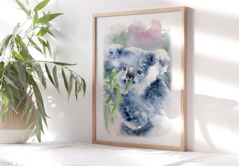 May include: Watercolour painting of a koala bear eating eucalyptus leaves. The koala is depicted in shades of blue and grey, with green leaves and a soft pink and green background. The artwork is framed in a light wood frame.