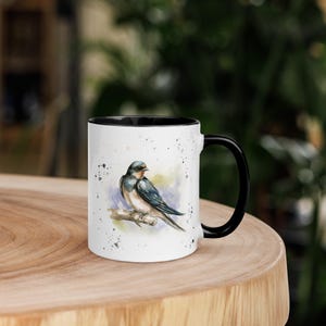 May include: White ceramic mug with a black rim and handle. The mug features a watercolor illustration of a swallow perched on a branch. The background is a light blue with white splatters.