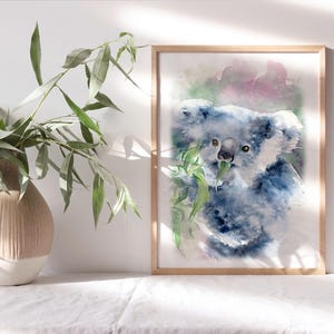 May include: A framed watercolour print of a koala bear, in shades of blue and grey, eating leaves. The frame is light wood. A vase with green leaves is to the left of the print.