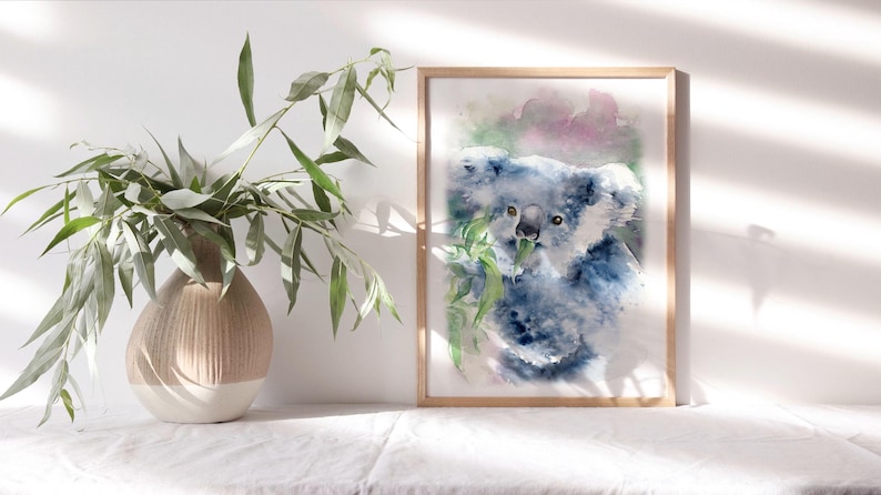 May include: A framed watercolour print of a koala bear, in shades of blue and grey, eating leaves. The frame is light wood. A vase with green leaves is to the left of the print.