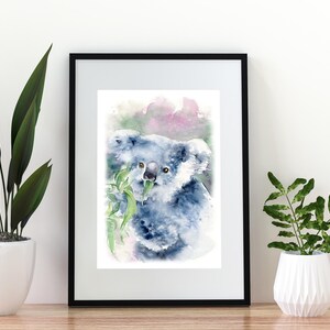 May include: Watercolour print of a koala bear eating eucalyptus leaves, framed in black. The artwork features soft blue and grey tones with touches of pink and green. The print is displayed on a wooden surface with potted plants on either side.