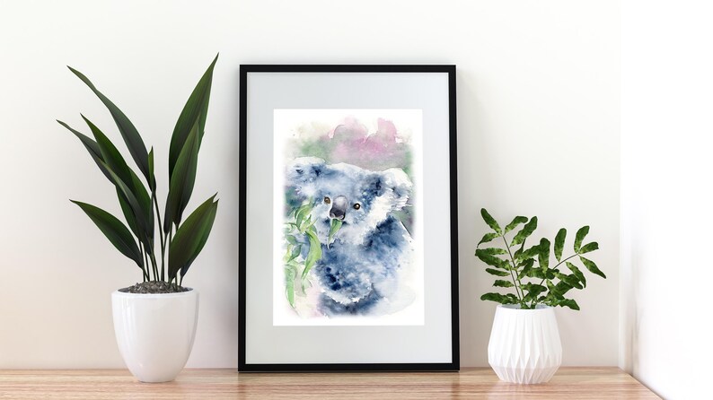 May include: Watercolour print of a koala bear eating eucalyptus leaves, framed in black. The artwork features soft blue and grey tones with touches of pink and green. The print is displayed on a wooden surface with potted plants on either side.