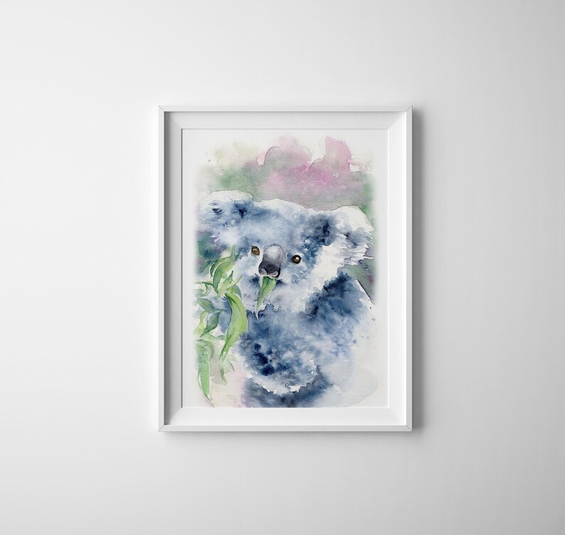 May include: A framed watercolour painting of a koala bear eating leaves. The koala is painted in shades of blue and grey, with green leaves and a soft pink and green background. The artwork is a vertical rectangle, framed in white.