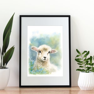 May include: Watercolor painting of a white lamb with brown eyes, standing in a field of blue flowers. The painting is framed in a black frame and is hanging on a white wall.