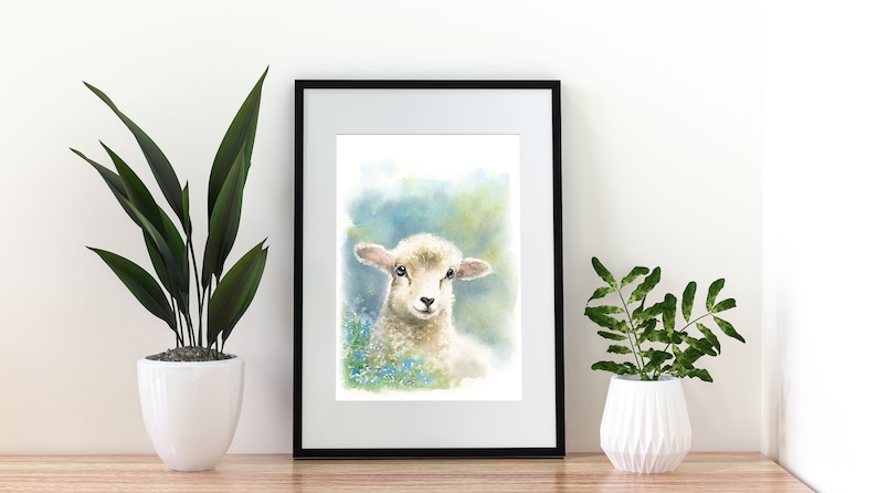 May include: Watercolor painting of a white lamb with brown eyes, standing in a field of blue flowers. The painting is framed in a black frame and is hanging on a white wall.