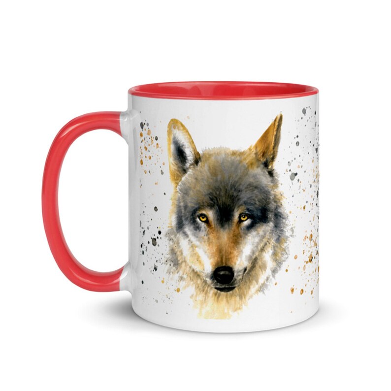 K&ouml;nnte beinhalten: Wei&szlig;e Keramiktasse mit rotem Henkel und Innenraum. Die Tasse zeigt ein Aquarellgem&auml;lde eines Wolfsgesichts mit gelben Augen. Spritzer in Braun und Schwarz akzentuieren das Design.