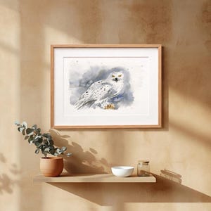 Könnte beinhalten: Ein gerahmtes Aquarellbild einer weißen Schnee-Eule mit schwarzen Punkten und gelben Augen vor einem weichen grau-blauen Hintergrund. Das Kunstwerk befindet sich in einem hellen Holzrahmen mit einem weißen Passepartout.