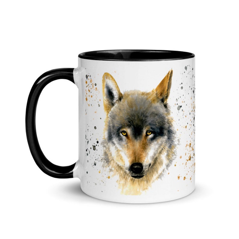 K&ouml;nnte beinhalten: Wei&szlig;e Keramiktasse mit schwarzem Innenraum und Griff. Die Tasse zeigt ein Aquarellgem&auml;lde eines Wolfsgesichts mit gelben Augen. Das Design umfasst braune und goldfarbene Farbspritzer.