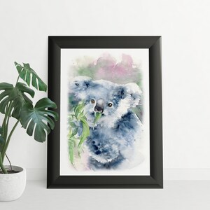 May include: Watercolour painting of a koala bear eating eucalyptus leaves, framed in a black frame. The artwork features shades of blue, grey, green, and pink. The koala is the central focus, with soft brushstrokes and a naturalistic style.