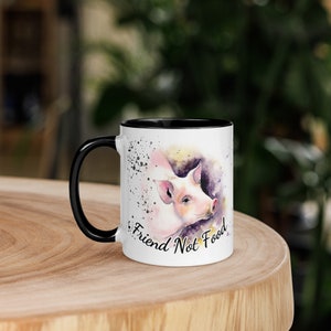May include: White ceramic mug with a black rim, featuring a watercolor illustration of a pig's face with the text "Friend Not Food".