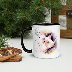 May include: White ceramic mug with a black rim and handle. The mug features a watercolor illustration of a pink pig with purple and yellow accents.