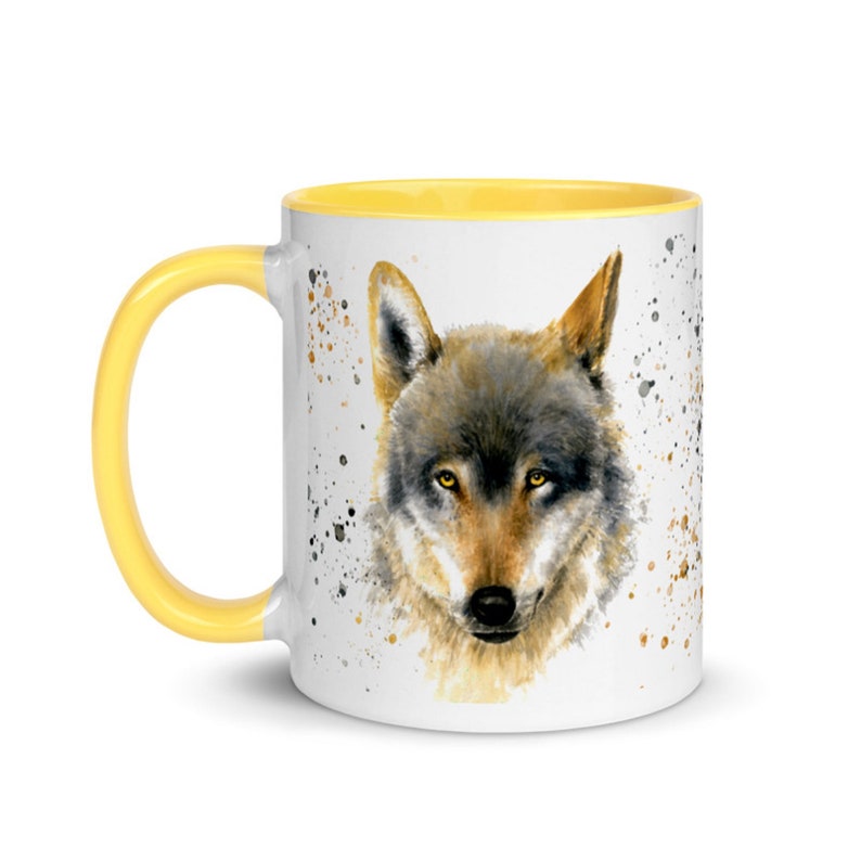 K&ouml;nnte beinhalten: Wei&szlig;e Keramiktasse mit gelbem Innenraum und Griff. Die Tasse zeigt ein Aquarellgem&auml;lde eines Wolfsgesichts mit Braun-, Grau- und Gelbt&ouml;nen. Spritzerartige Farbflecken in Braun und Schwarz umgeben den Wolf.