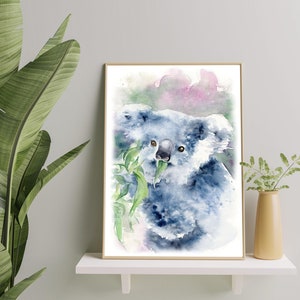 May include: Watercolour painting of a koala bear eating leaves, framed in a gold border. The koala is in shades of blue and grey, with green leaves. The background features soft pink and green hues. The artwork is displayed on a white shelf.