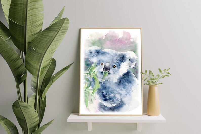 May include: Watercolour painting of a koala bear eating leaves, framed in a gold border. The koala is in shades of blue and grey, with green leaves. The background features soft pink and green hues. The artwork is displayed on a white shelf.
