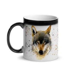 K&ouml;nnte beinhalten: Eine wei&szlig;e Keramik-Tasse mit schwarzem Henkel und Rand. Die Tasse zeigt ein Aquarellgem&auml;lde eines Wolfsgesichts mit braunen, grauen und goldenen Farbt&ouml;nen. Spritzer in Braun und Gold akzentuieren das Design.