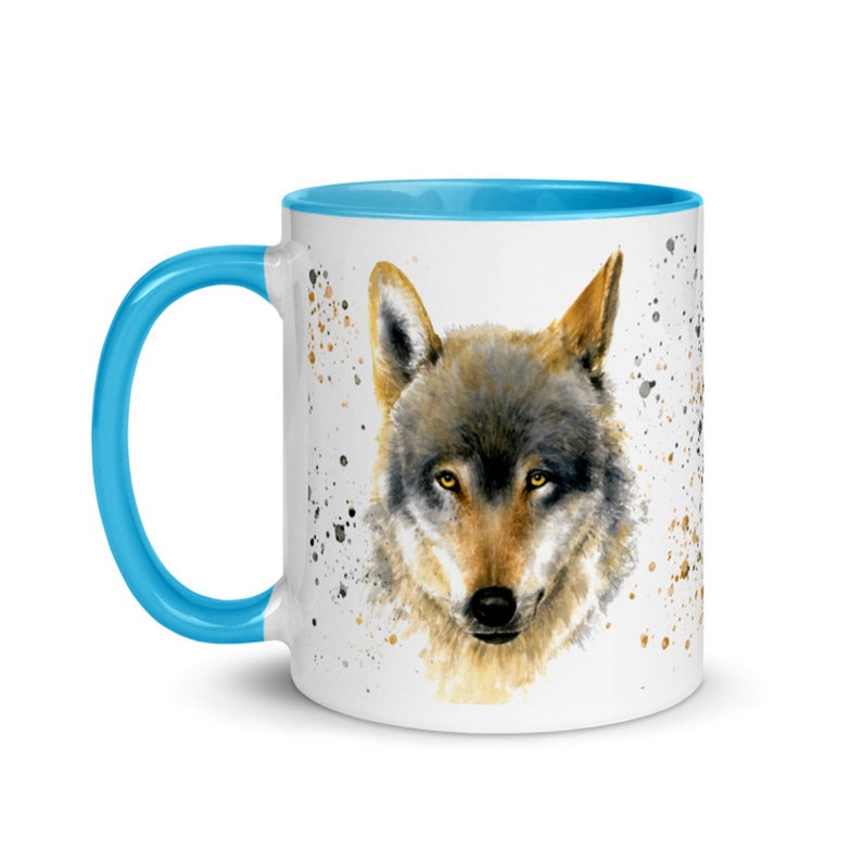 K&ouml;nnte beinhalten: Wei&szlig;e Keramiktasse mit einer Aquarellillustration eines Wolfsgesichts. Die Tasse hat einen hellblauen Innenraum und Henkel mit braunen und schwarzen Farbspritzern. Der Wolf hat gelbe Augen und graues und braunes Fell.