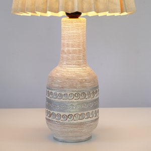 May include: A white ceramic table lamp with a pleated fabric lampshade. The lamp base has a textured, woven design and a silver-coloured band with a repeating pattern.