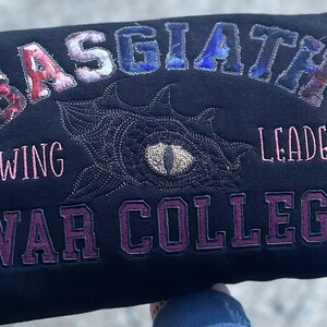 May include: Black sweatshirt featuring embroidered text and a dragon eye design. The text reads "BASGIATH WING LEADER WAR COLLEGE" in red, blue, and purple. The dragon eye is in the centre.