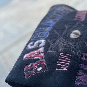 May include: Dark fabric with embroidered lettering and a floral patch. The text includes "BASIL" in a red floral pattern, "WING" in pink, and "WAR COLLEGE" in purple. An embroidered eye design is also visible.