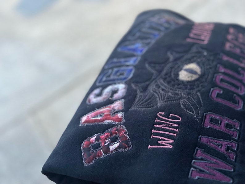May include: Dark fabric with embroidered lettering and a floral patch. The text includes "BASIL" in a red floral pattern, "WING" in pink, and "WAR COLLEGE" in purple. An embroidered eye design is also visible.