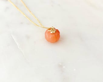 May include: A gold chain necklace with a small, orange-colored pumpkin pendant. The pumpkin pendant has a gold-colored cap and is carved with detailed ridges. The necklace is displayed on a white marble surface.