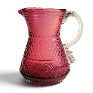 May include: A translucent cranberry-colored glass pitcher featuring a clear glass handle. The body displays a textured pattern with a diamond-shaped grid on the lower section and a subtle swirling design on the neck, finished with a flared rim.