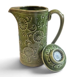 May include: A tall, cylindrical green ceramic pitcher with a handle and a matching circular lid. The exterior features a raised, embossed floral pattern with chrysanthemum-like motifs and swirling vines, finished with a glossy, crackled glaze.