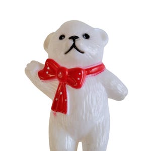 May include: A white teddy bear figurine with a red bow tie featuring white polka dots. The bear has black eyes and a black nose. The bear is waving its left arm. The figurine is made of a smooth, glossy material.