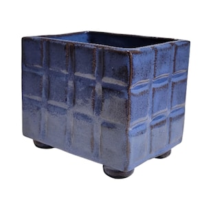 May include: A square, ceramic planter in a deep blue hue. The exterior features a textured, grid-like pattern. The planter rests on four small, rounded feet. The interior is a darker shade of blue.