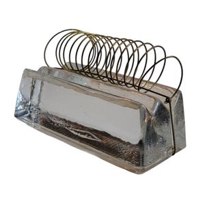 May include: A clear glass rectangular desk organizer with a brass wire spiral. The organizer has a thick, textured base and a series of brass wire loops for holding papers or notes. The design is simple and modern.
