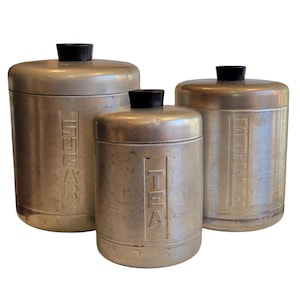 May include: Three vintage metal canisters with black lids. The largest canister is labeled "SUGAR", the middle one "TEA", and the smallest "SUGAR". The canisters have a brushed metal finish and are likely from the mid-20th century.