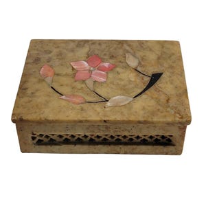 May include: A rectangular, beige stone box with a floral design on the lid. The flower is pink with a mother-of-pearl center, and the leaves are white. The box has a decorative, geometric pattern along the front edge.
