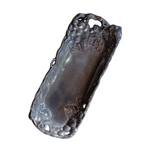 May include: A rectangular, silver-toned metal tray with a handle. The tray is decorated with grape clusters and leaves along the edges. The handle is arched and also features grape details. The tray is empty.