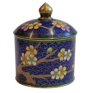 May include: A cylindrical metal jar with a removable lid featuring a pointed finial. The exterior is decorated with blue enamel, a repeating cloud pattern, and branches with yellow and white flowers. The rim and base have green scalloped borders.
