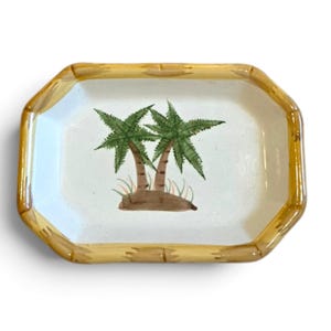 May include: A rectangular ceramic tray with a white center and a yellow border painted to resemble bamboo segments. The center features a hand-painted illustration of two green palm trees growing from a small brown mound of earth with grass.