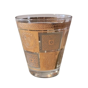 May include: A clear glass tumbler with a gold and silver geometric design. The design includes a sun, squares, and a lyre. The tumbler has a wide mouth and a slightly tapered shape, suitable for serving beverages.