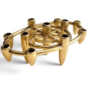 May include: A gold-toned metal candelabra featuring an oval-shaped frame with multiple candle holders. The design includes a central wheel-like structure with spokes connecting to the outer ring, supported by pointed, tapered feet at each holder.