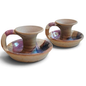 May include: Two ceramic candle holders with handles, featuring a speckled tan base color and glazed accents in shades of purple, blue, and brown. Each holder has a wide, circular drip tray and a central cylindrical socket for a taper candle.