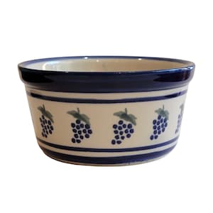 May include: A ceramic bowl with a cream-colored base and a blue rim and stripes. The bowl is decorated with clusters of blue grapes and green leaves. The bowl is a round shape.