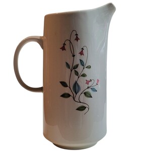 May include: White ceramic pitcher with a curved handle and spout. The pitcher is decorated with a floral design featuring pink bell-shaped flowers, green leaves, and blue stems. The pitcher is approximately 8 inches tall.