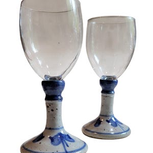 May include: Two wine glasses with clear bowls and ceramic bases. The bases are speckled with a blue and white floral design. The stems are a solid blue color. The glasses are on a white background.