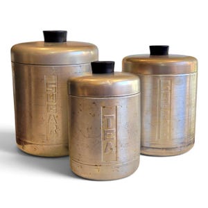 May include: A set of three cylindrical brushed metal canisters with black plastic knobs on the lids. Each canister features vertical embossed lettering on the front, specifically labeled as SUGAR, TEA, and COFFEE, with a weathered metallic finish.