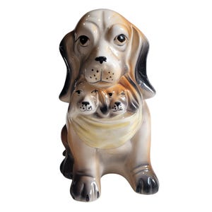 May include: A ceramic figurine depicting a large dog with long, dark-tipped ears and a light brown coat, featuring two smaller puppies nestled in front of its chest. The puppies are wearing a smooth, pale yellow neckerchief around their necks.